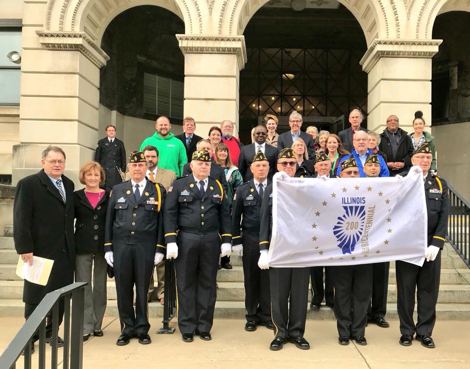Members of the public, elected officials, and the local American Legion Honor Guard/Color Guard pose with the Illinois Bicentennial flag before the ceremony on Monday, Dec. 4, 2017. (Photo by Candace Summers)