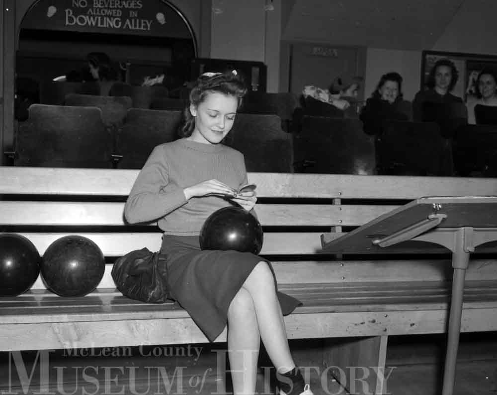 Photo of a woman sitting on a bench with a bowling ball on her lap.