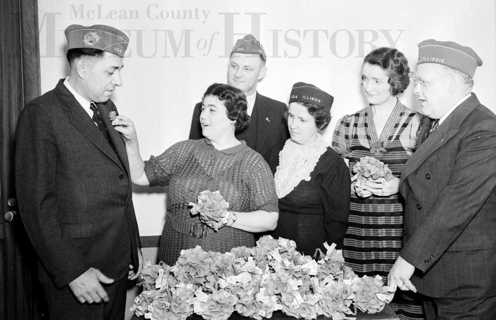 Three men and three women are pictured with a pile of flowers in the foreground. One of the women is pinning a flower to one of the Men's jackets, while the others watch.